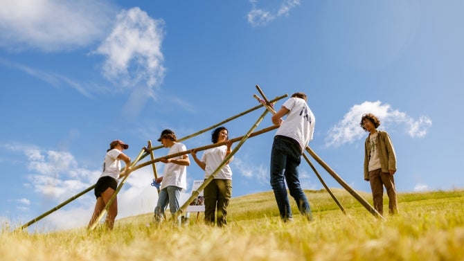 A group of students assembling bamboo poles in a grassy field under a bright blue sky.