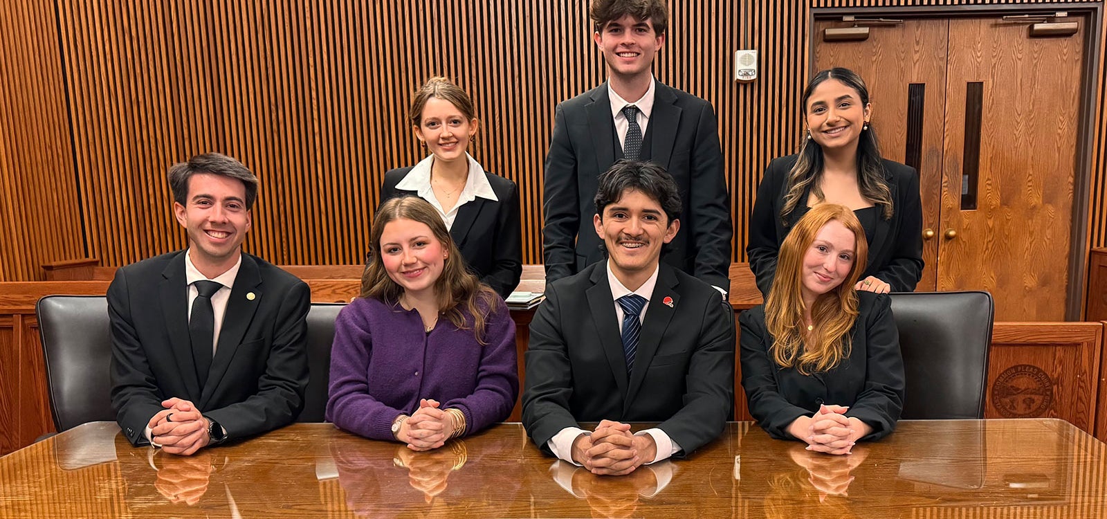 Group of mock trail students sitting next to a wooden table dress in formal business attire.