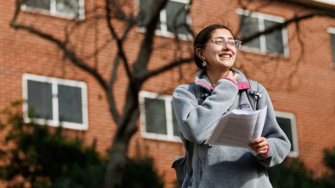 Smiling student holding papers outside a brick campus building on a sunny day.