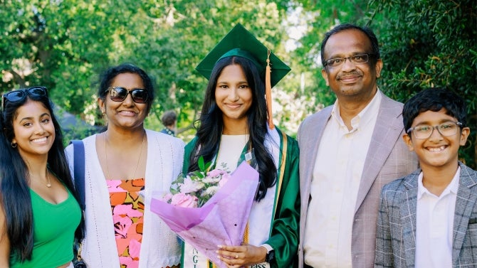 Graduate in green cap and gown holding flowers, celebrating with family outdoors.