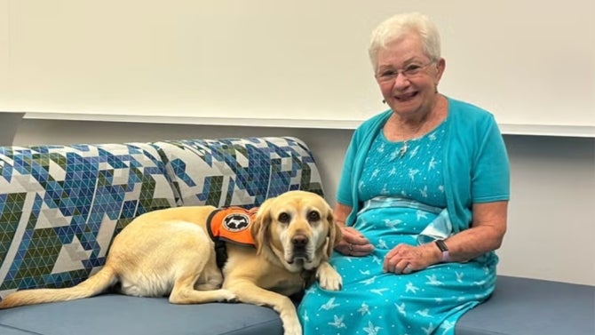Noni Smyth in turquoise outfit sitting on couch with yellow Labrador service dog wearing orange vest.