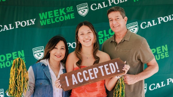 Student holding 'I ACCEPTED!' sign with parents at Cal Poly welcome event.