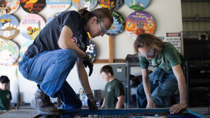 Students in safety glasses working on a metal project in a workshop, with colorful circular artwork displayed on the wall behind them.