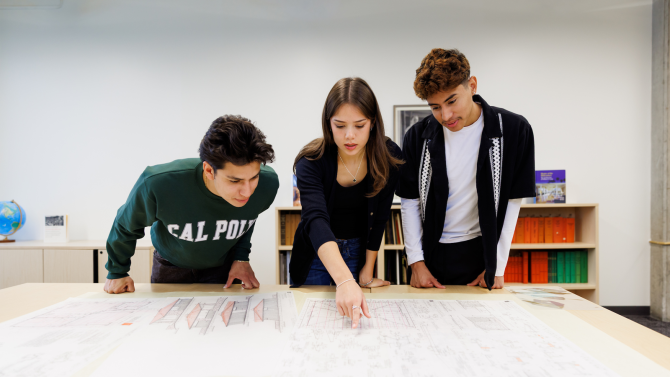 3 people hover over a table to read drawings on a paper